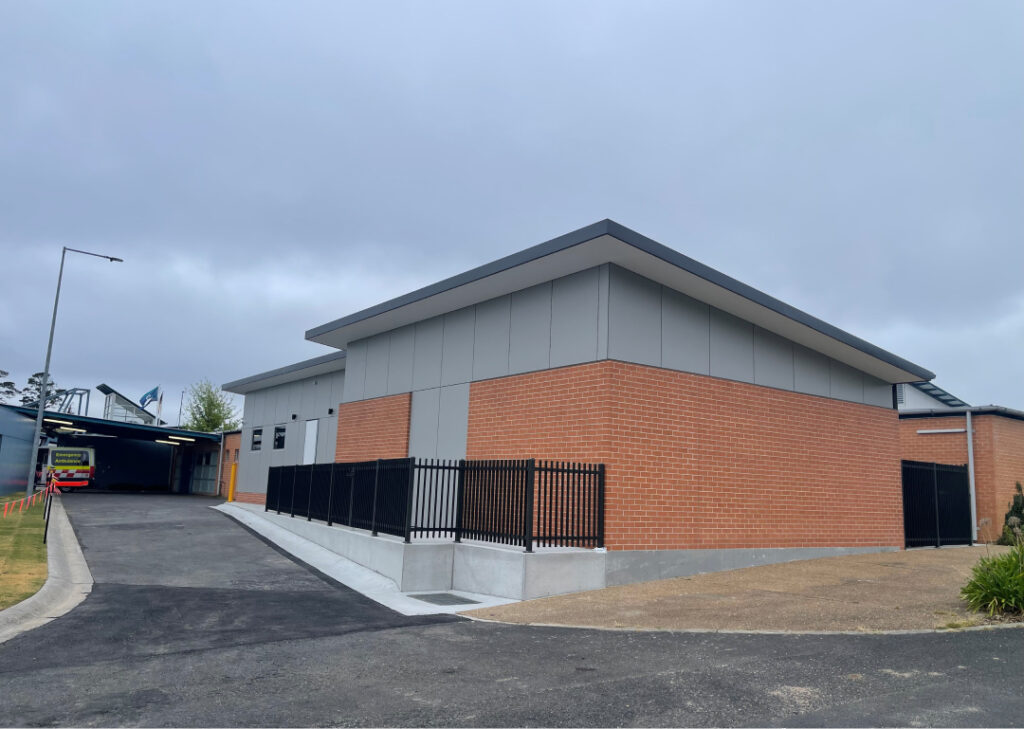 A modern, single-story building features a mix of brick and gray siding. A black metal fence encloses the side. The driveway next to the building extends to a covered area with a yellow vehicle partially visible. The sky is cloudy and overcast.