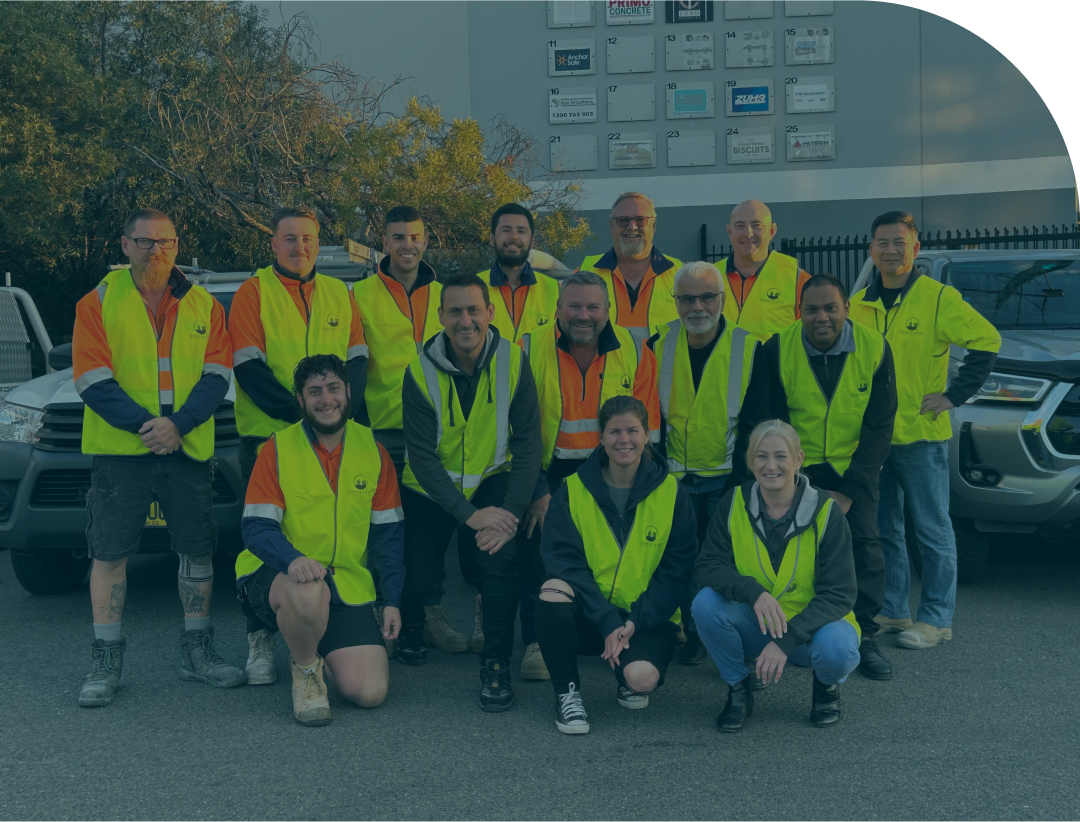 A group of fourteen people in high-visibility jackets pose for a photo in front of two vehicles, showcasing their Enginuity spirit. The backdrop includes a building with multiple company signs, while trees and fencing complete the scene.