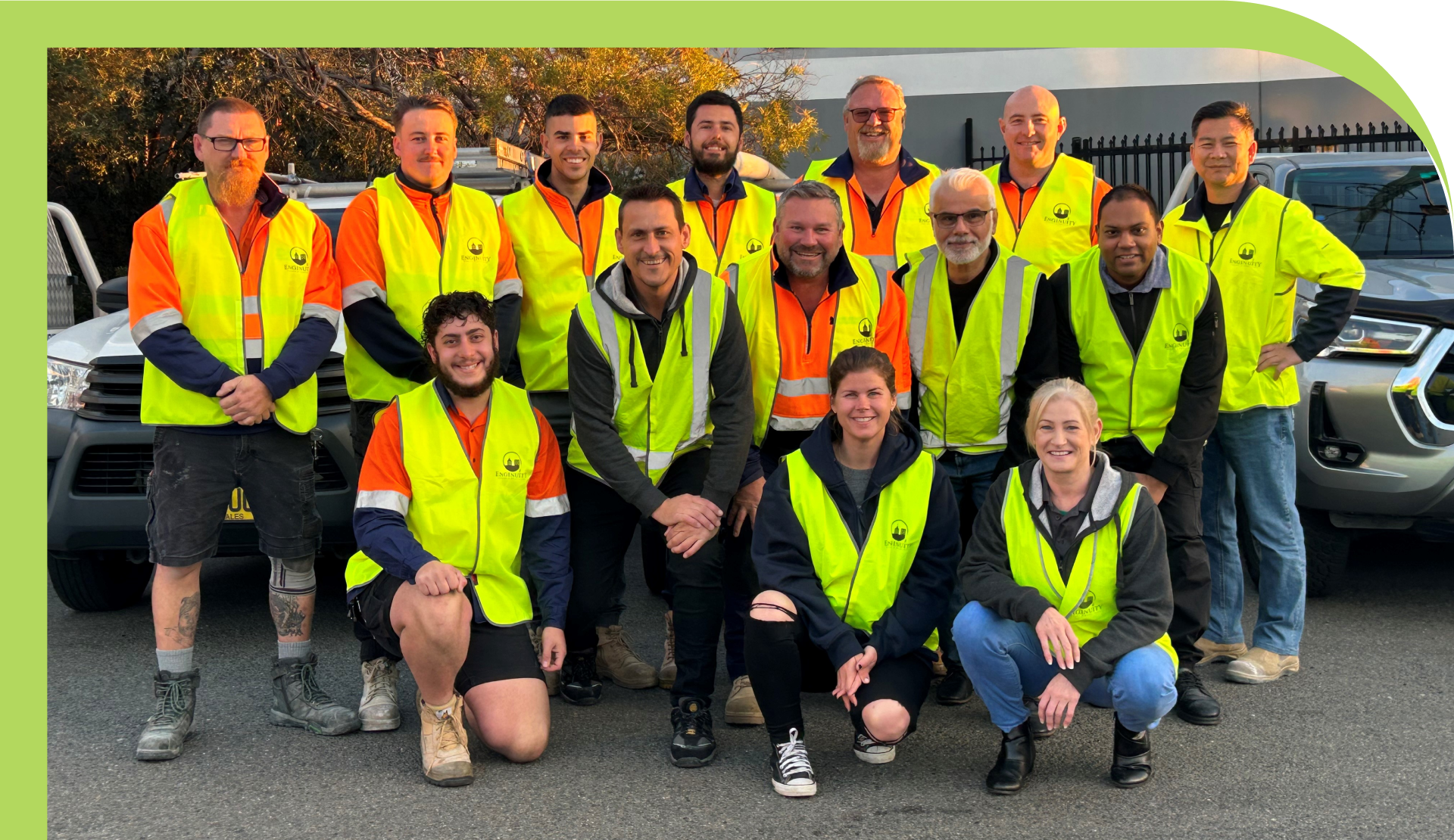 A group of twelve people, wearing high-visibility vests, pose for a photo outdoors. Some are standing while others are kneeling in front. They are in a parking area with vehicles and a fence visible in the background, symbolizing the teamwork and dedication that embodies About Enginuity.