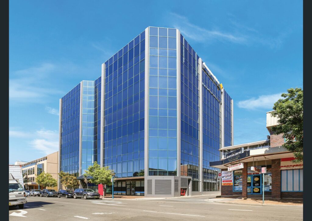 A modern, blue-glass commercial building stands on a sunny street corner, with a few cars parked along the curb. The structure features reflective windows and adjacent lower buildings, and the sky is clear and blue. Signs and trees are visible at street level.