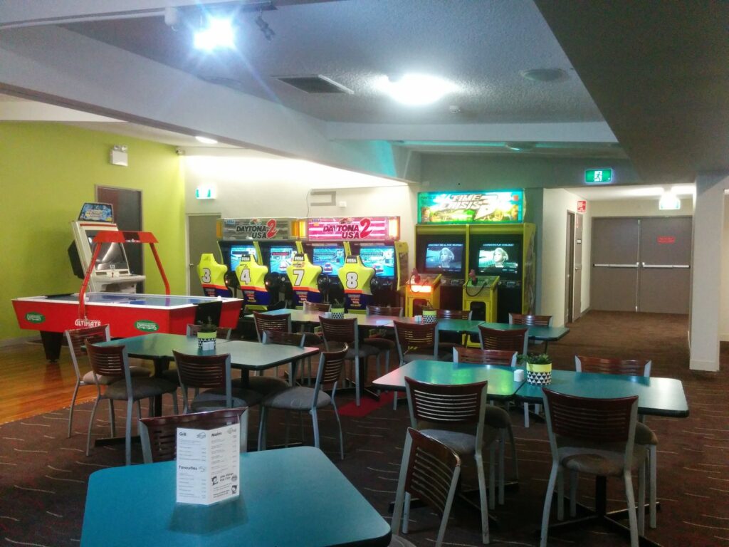 A room with several arcade game machines and an air hockey table. In front, there are tables and chairs arranged for seating. Walls are painted green and white, and the room is well-lit with overhead lights. Emergency exit signs are visible above doorways.