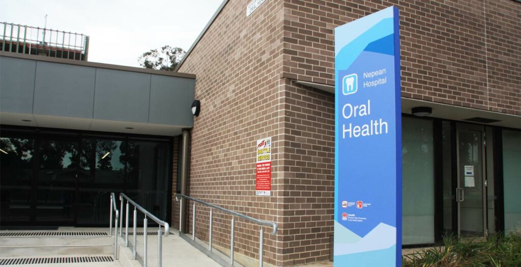 A brick building with a sign outside that reads "Napean Hospital Oral Health." The entrance features a handicap-accessible ramp and glass doors. Various informational posters are displayed on the wall near the entrance.