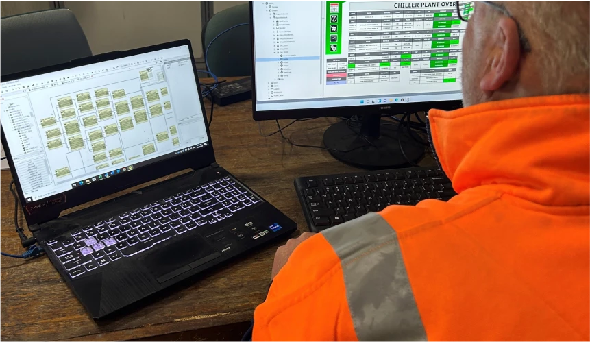 A person wearing an orange high-visibility jacket is working at a wooden desk with a laptop and a desktop monitor. The laptop shows a detailed flowchart, while the monitor displays a spreadsheet titled "Chiller Plant Overview." This scene reflects the meticulous attention to detail that defines Enginuity.